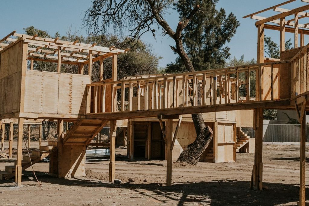 A sunlit rural scene of unfinished wooden structures with trees in the background.